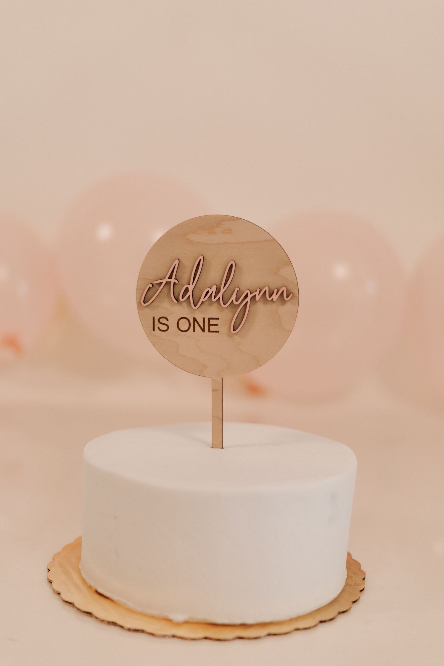 White frosted cake with wooden cake topper decorations on a light pink background.