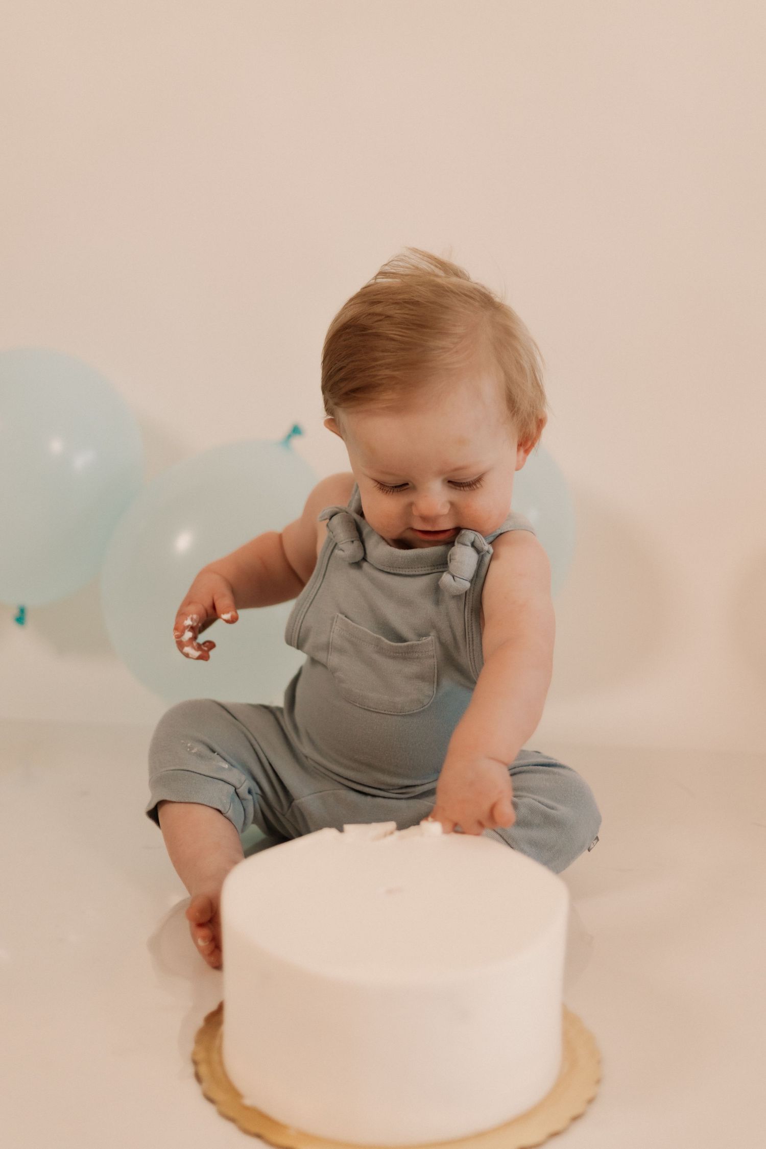 Baby in grey romper exploring a white birthday cake during a cake smash photoshoot.