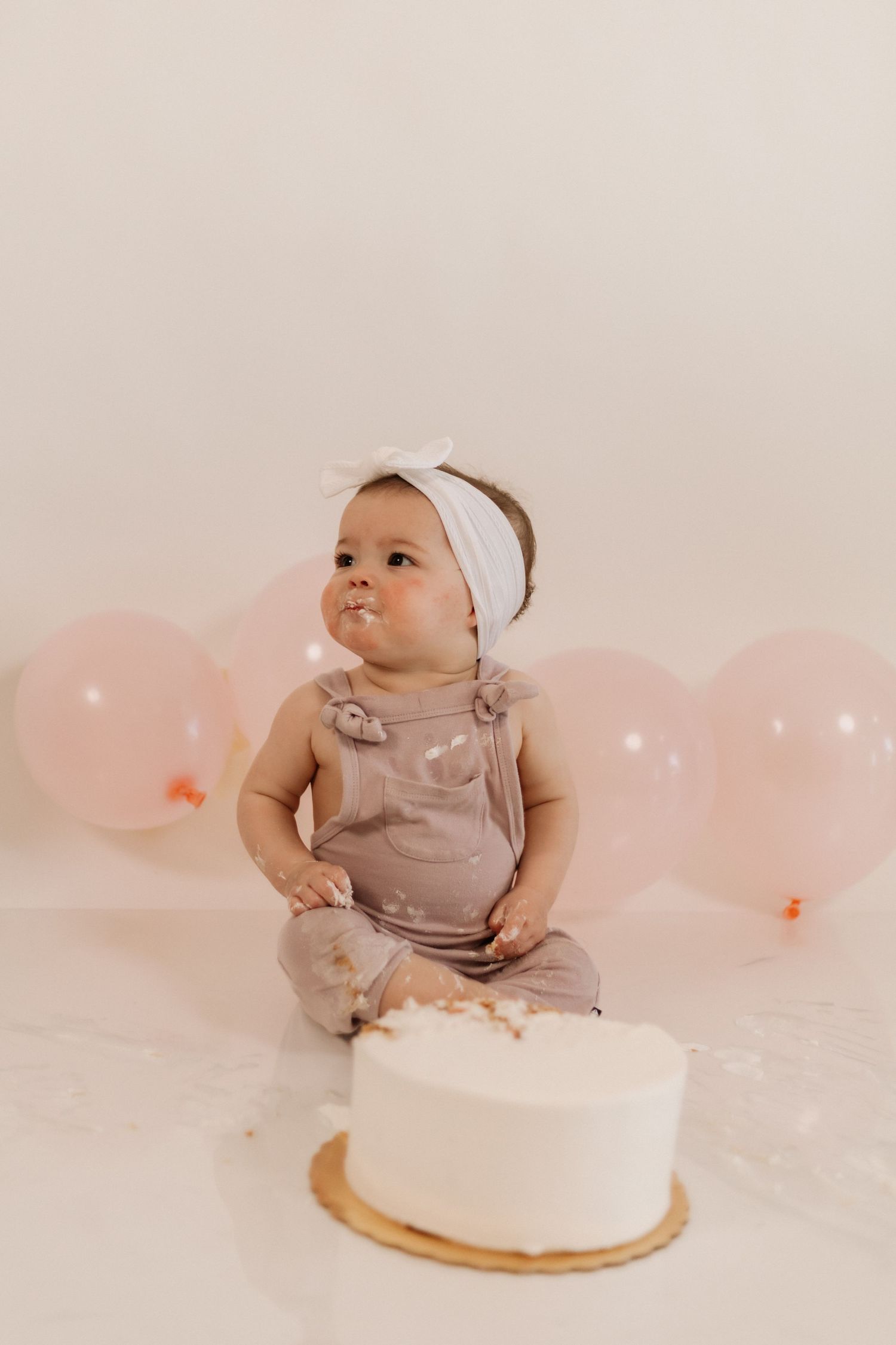 Baby in beige outfit sitting with white cake and pink balloons during first birthday photoshoot.