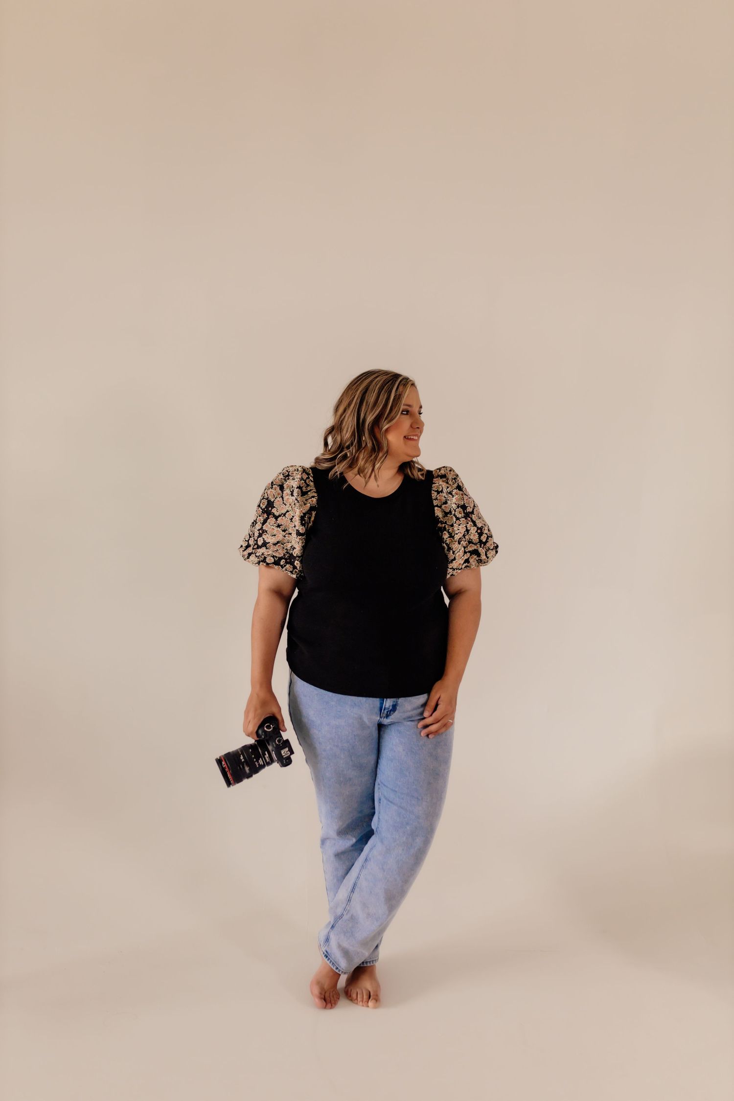 Woman in black top and jeans posing casually against white studio backdrop.