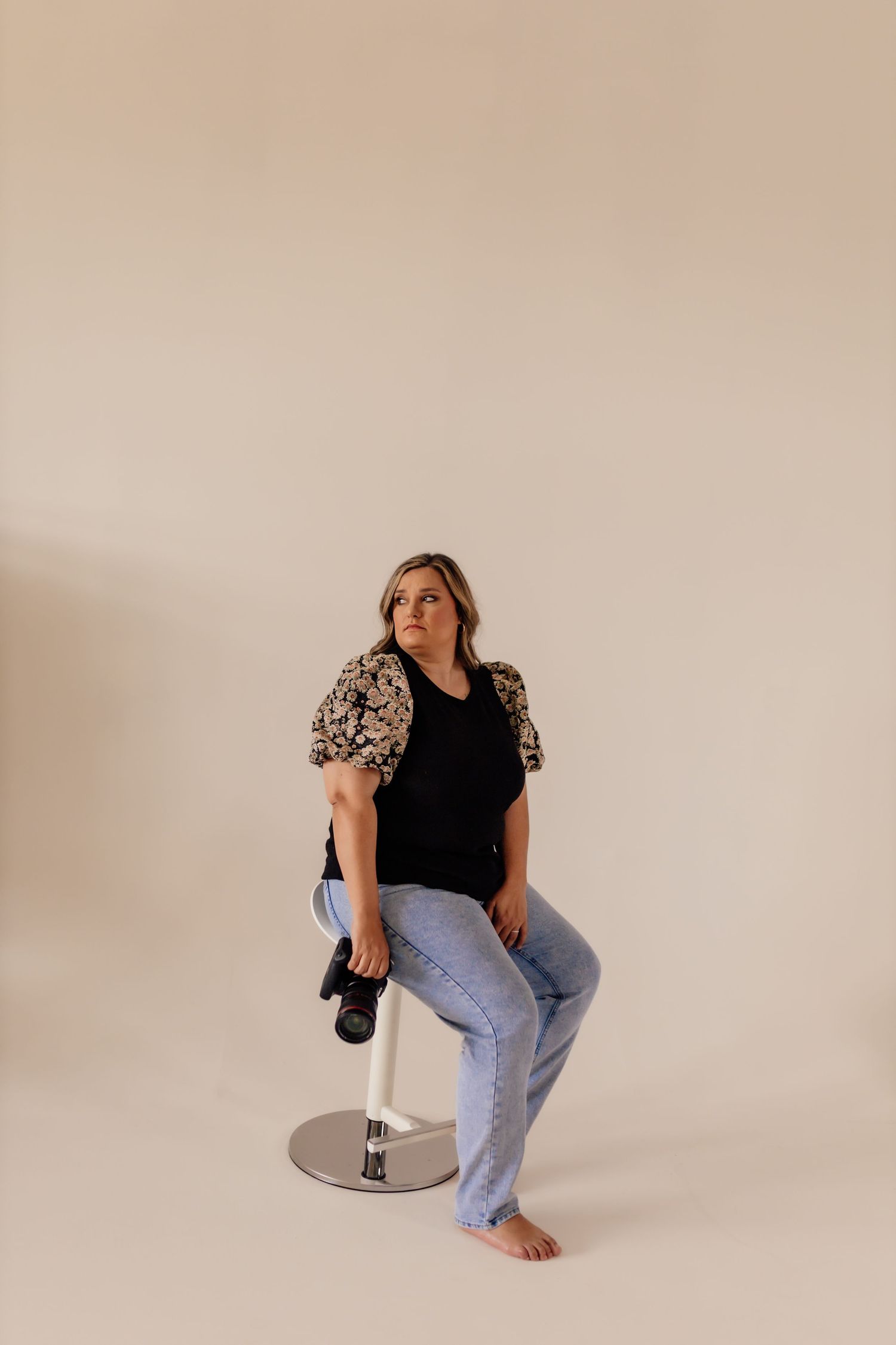 Woman in black top and jeans poses on silver stool against neutral background.