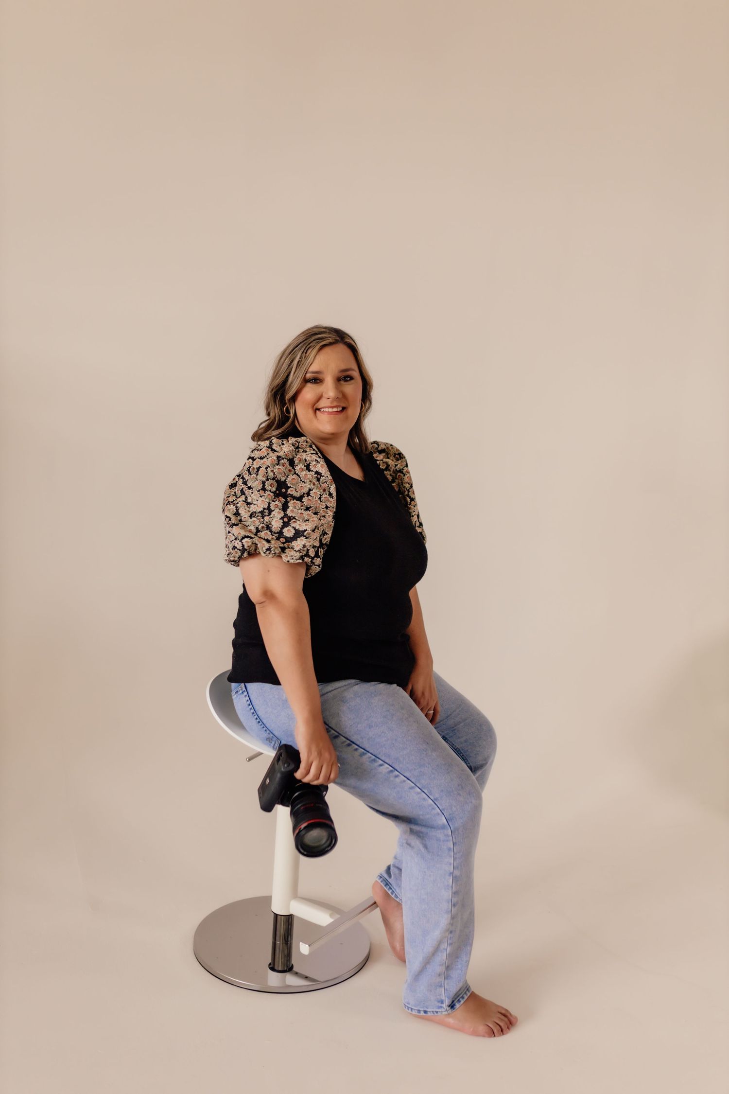 Woman in floral sleeve top and jeans sits casually on silver stool for portrait session.
