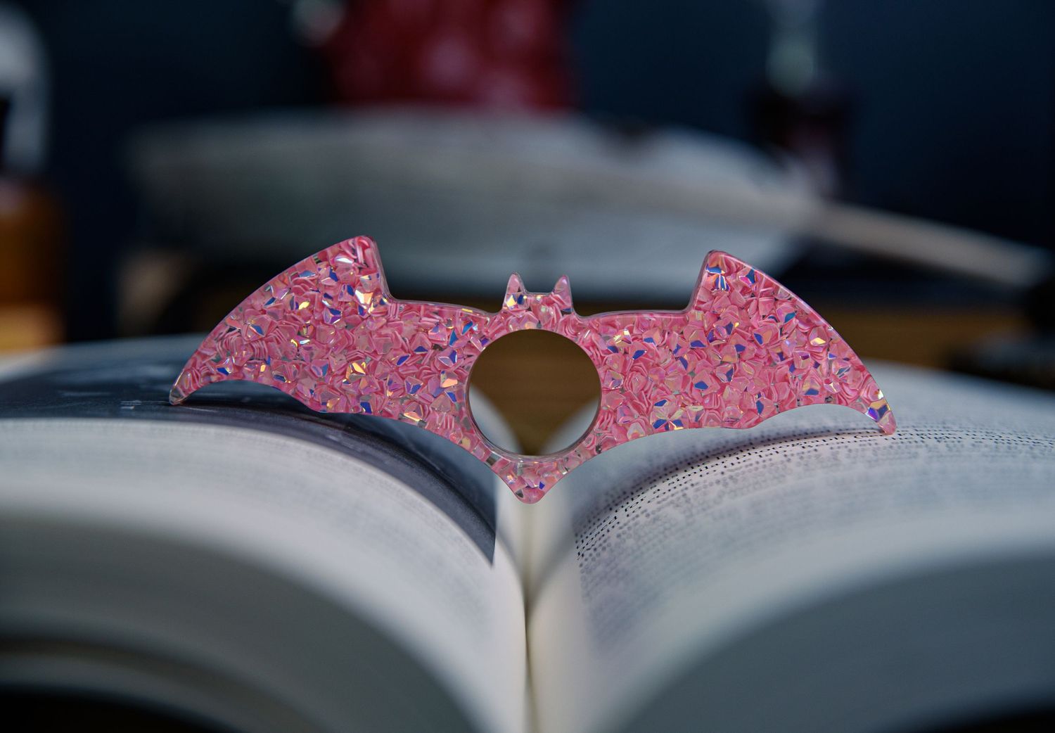 Pink resin book page holder, bat-shaped, sitting on an open book in a moody academic setting