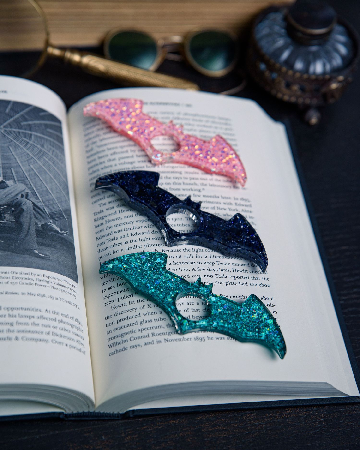 Resin book page holders, bat-shaped, in various colors lying on an open book in a moody academic setting