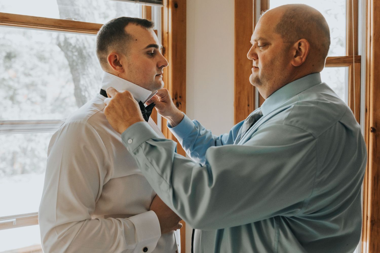 Two people in formal attire stand by a window as one adjusts the other's bow tie in an intimate getting-ready moment.