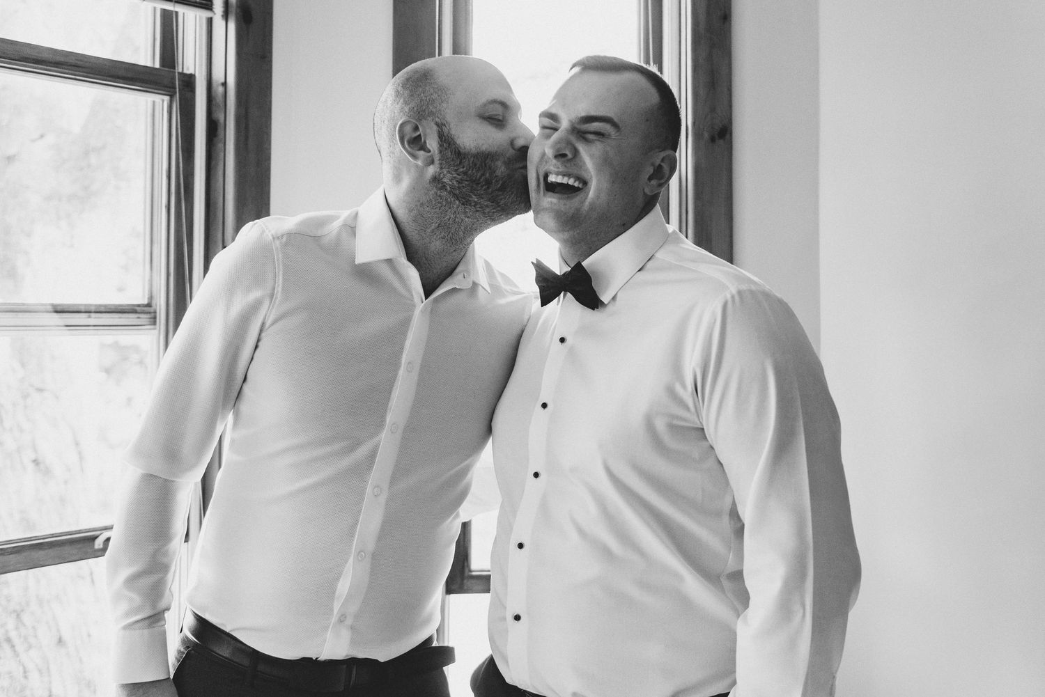 A tender moment of two men in white dress shirts laughing and embracing by a window in black and white photography.
