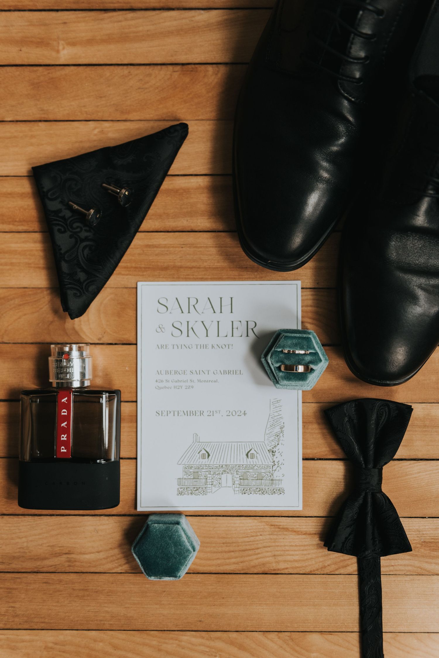 Wedding details flatlay with invitation, black dress shoes, bow tie, cologne bottle and emerald decorative elements on wooden floor.