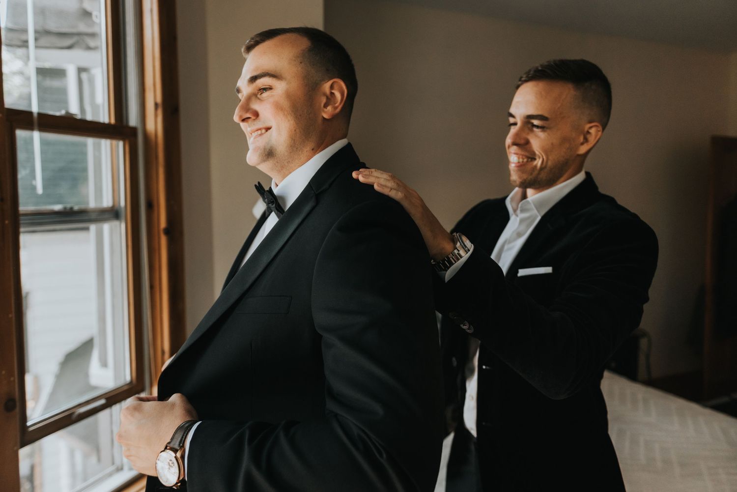 Two people in black suits stand by a window in a room, with one adjusting the others clothing.