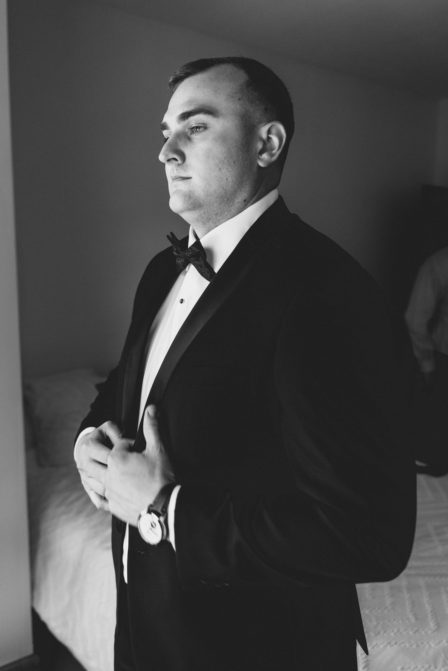 Black and white portrait of someone in a tuxedo adjusting their jacket while looking sideways in dramatic lighting.
