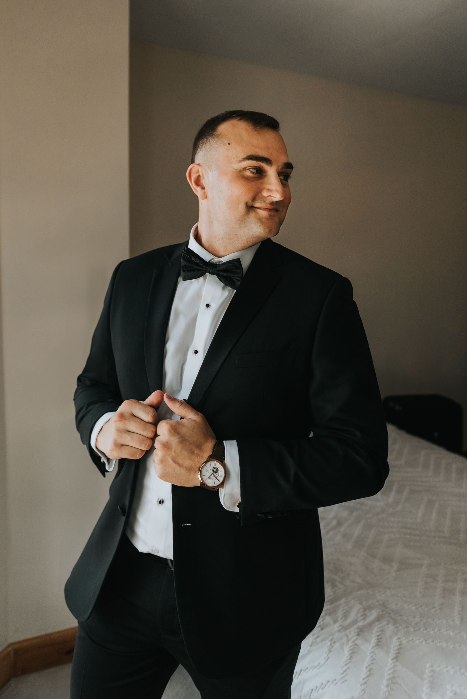 Someone in a classic black tuxedo and bowtie adjusts their formal wear while standing beside a bed.