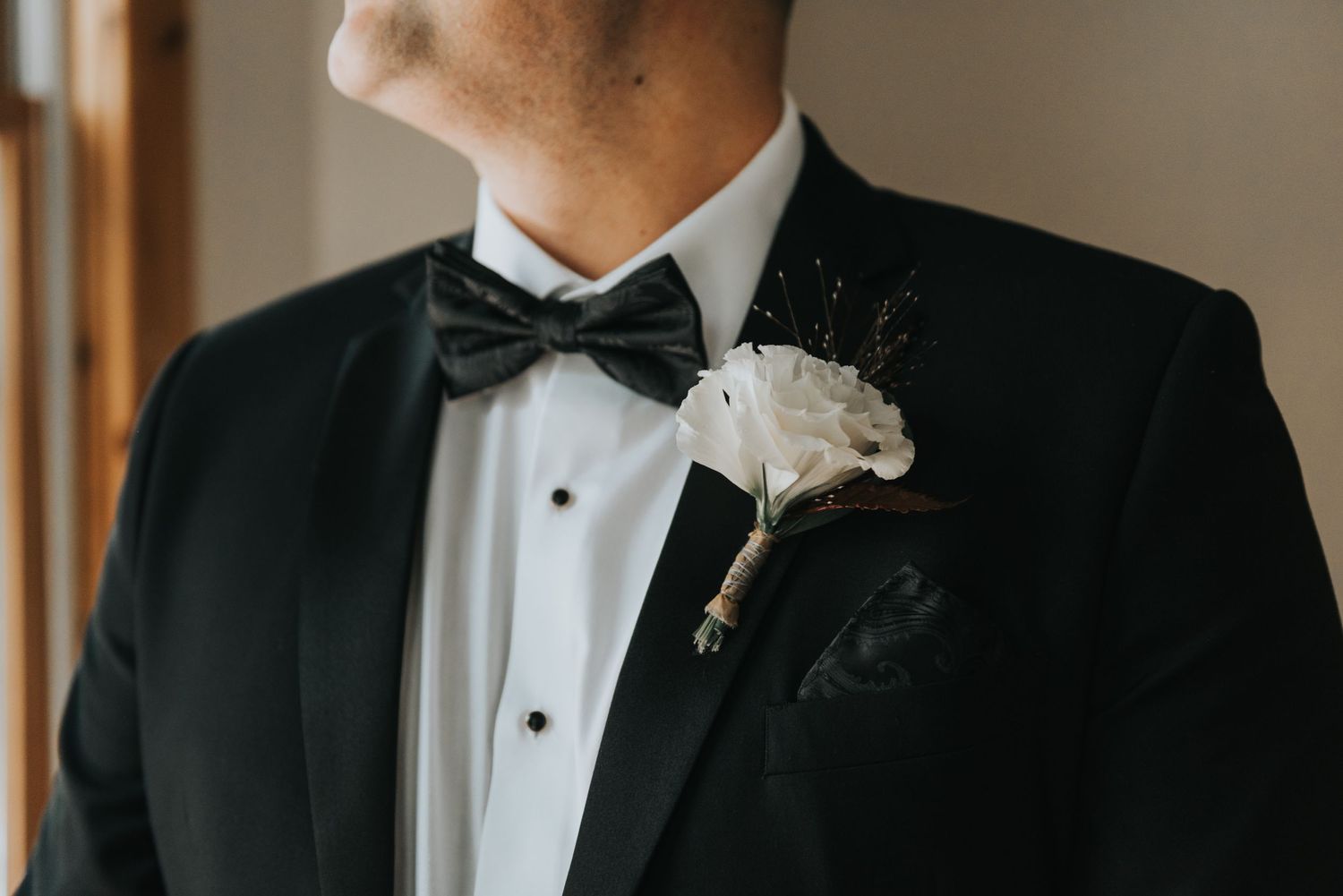 Close-up detail of black tuxedo with bow tie and white rose boutonniere against window light.