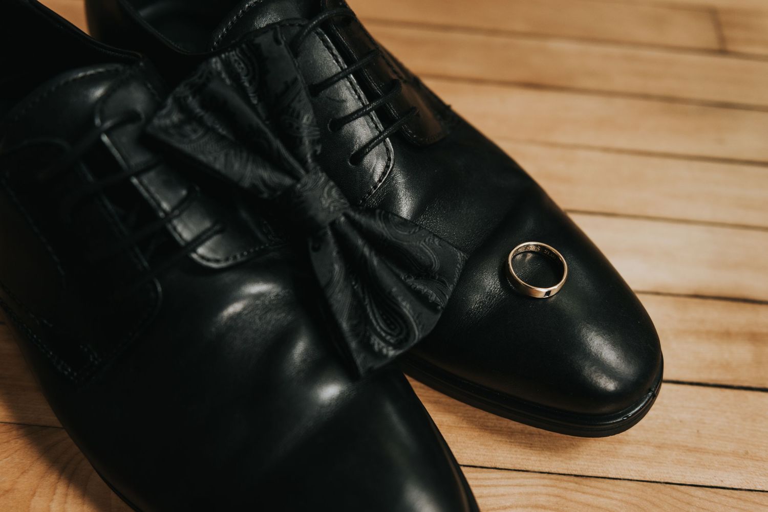 A black leather dress shoe with a decorative bow and button detail sits on a wooden floor.