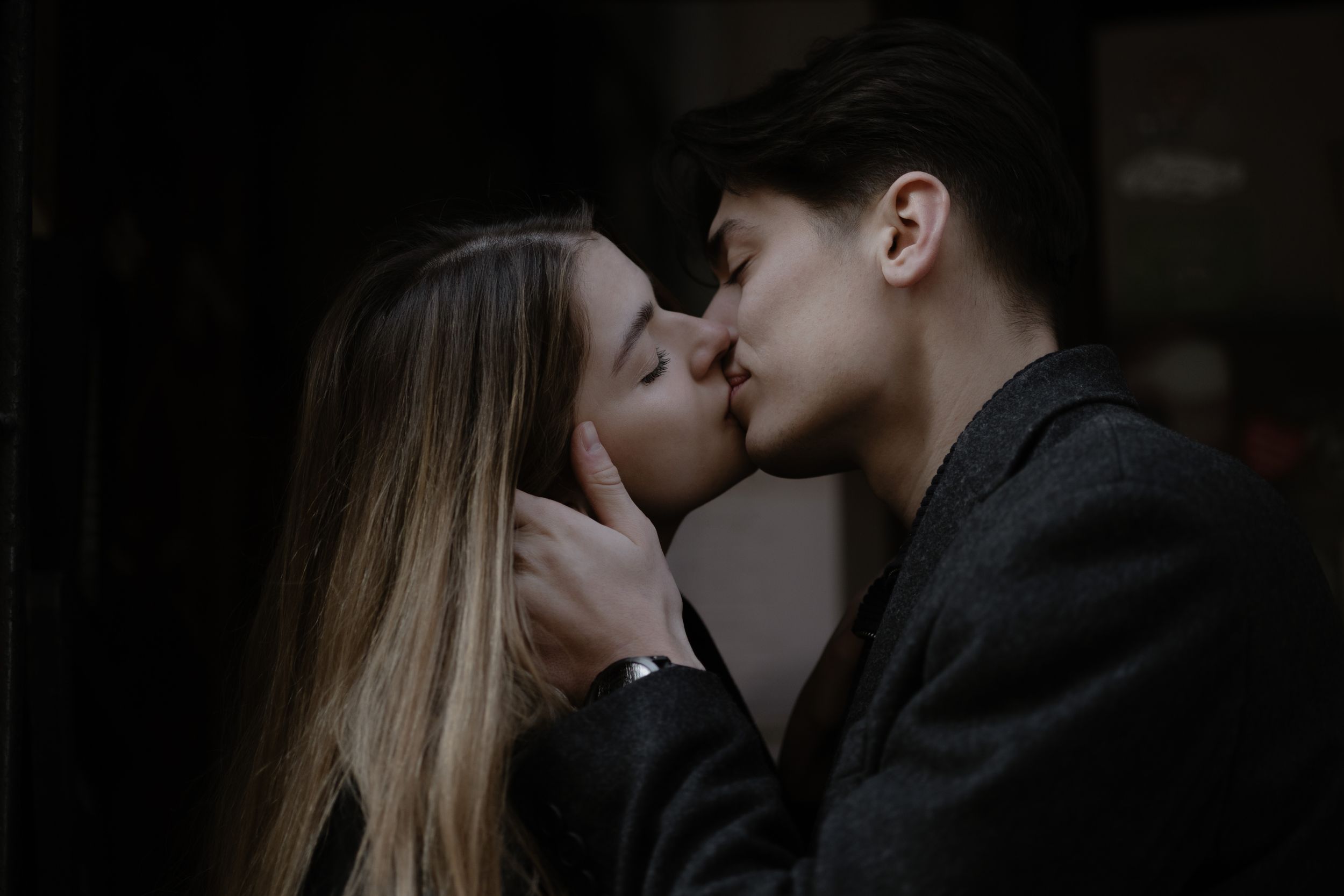 A loving couple shares a romantic kiss in a dark, intimate setting with soft atmospheric lighting.