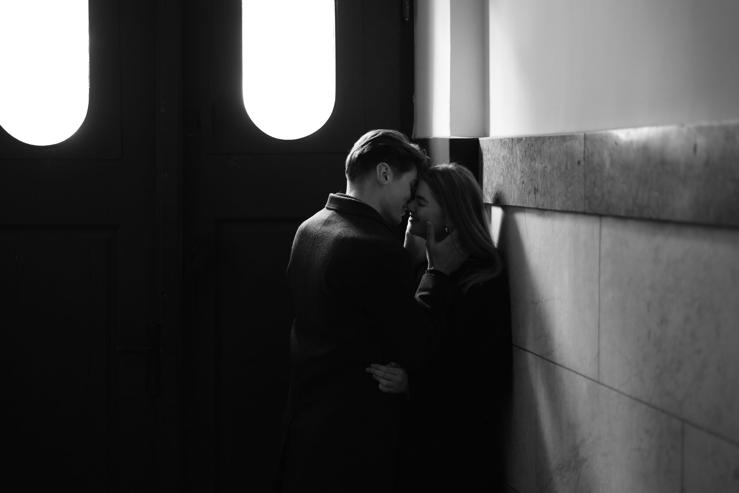 A moody black and white photograph of a couple sharing an intimate moment against a wall with oval windows.
