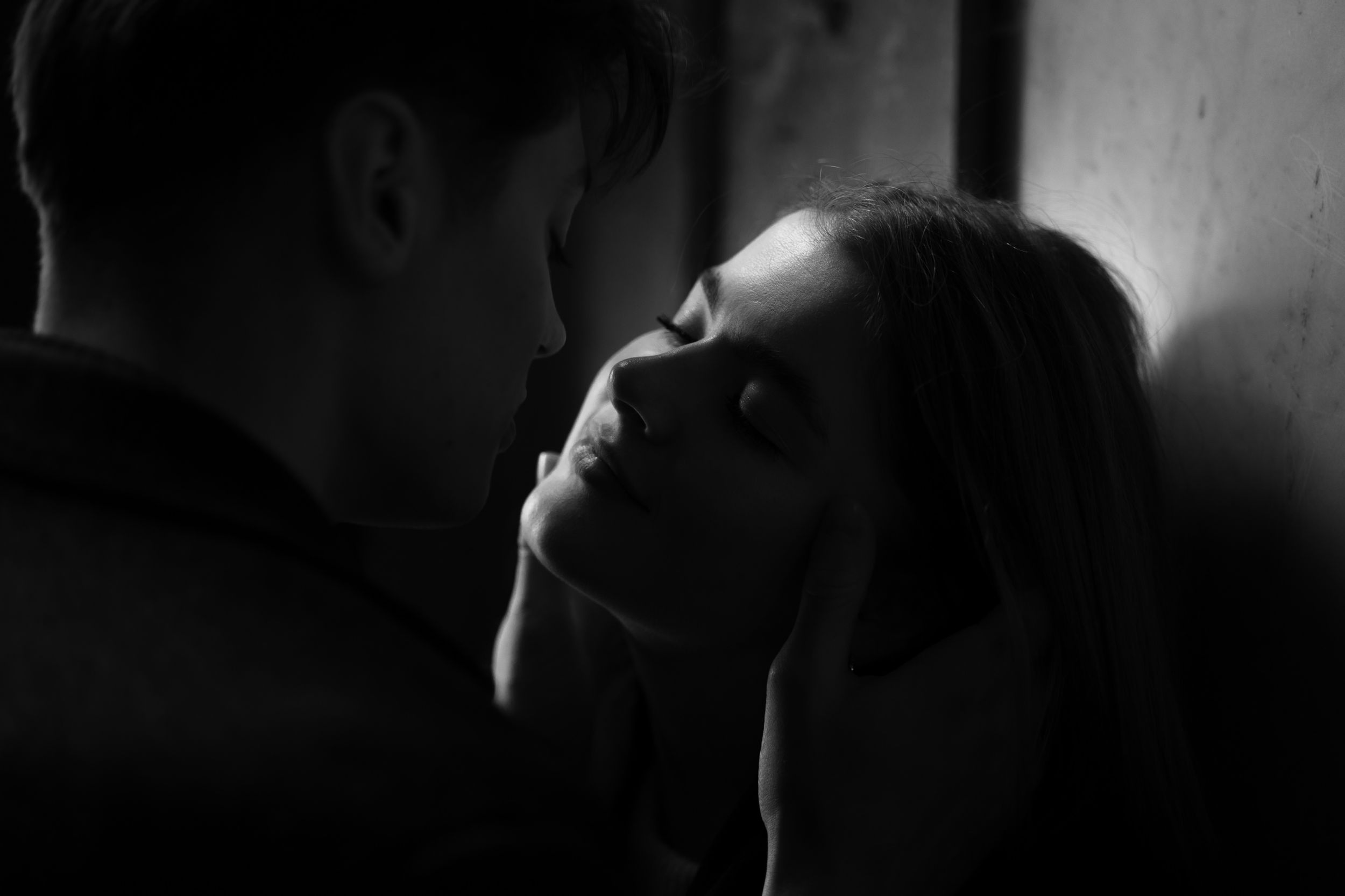 A dramatic black and white intimate portrait of two people in shadows with one leaning back against a wall.