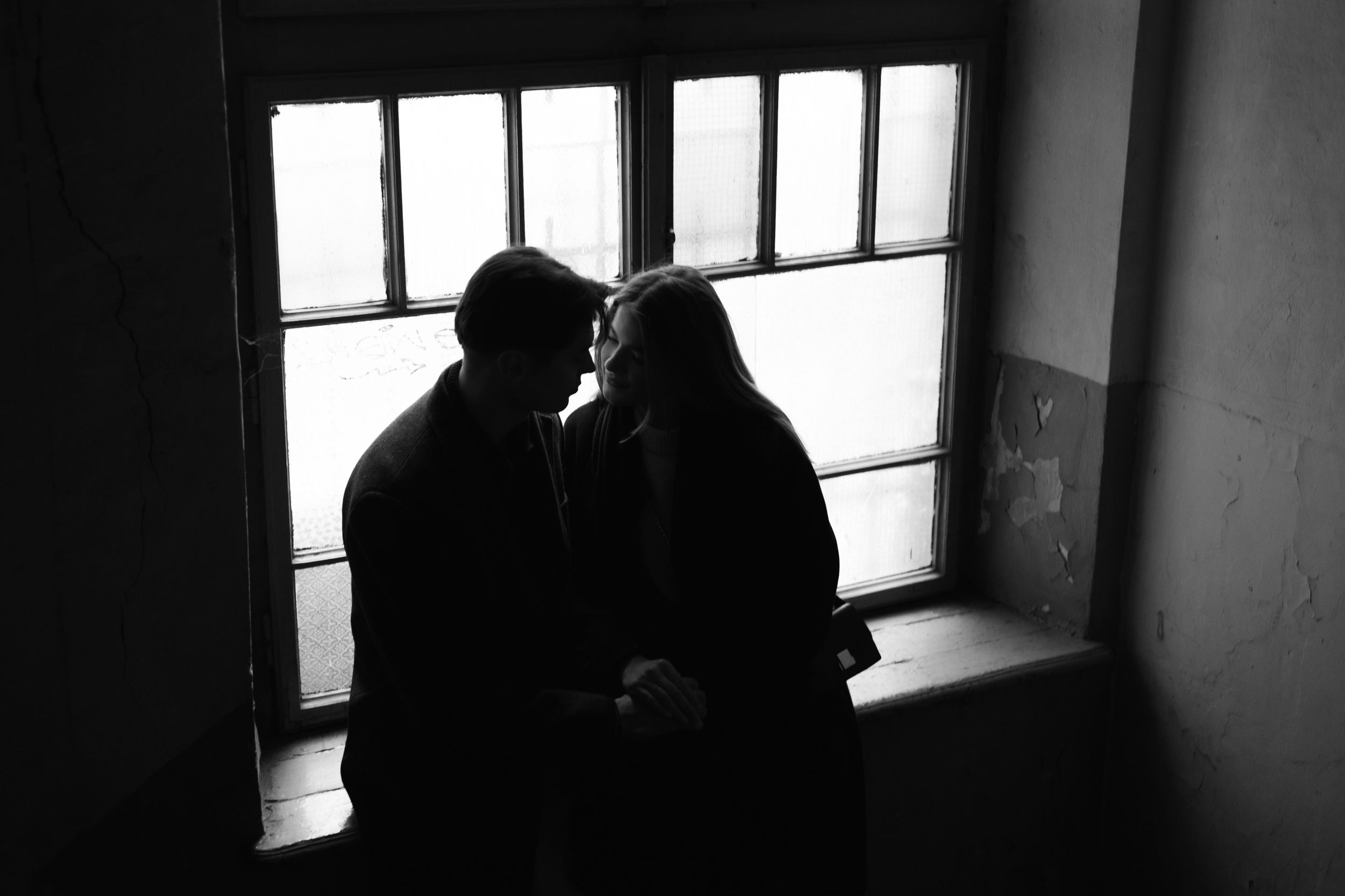 A silhouetted couple share an intimate moment by a bright window in a black and white photograph.