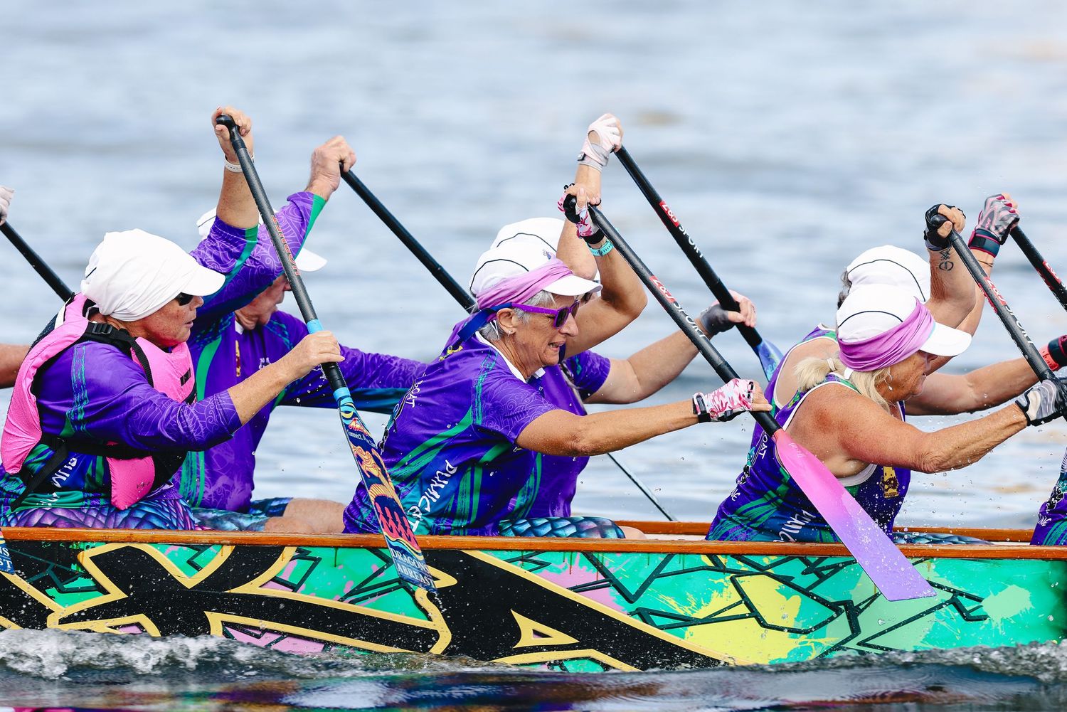 Dragon boat racers in purple and pink uniforms paddle in synchronized motion during a competitive race.