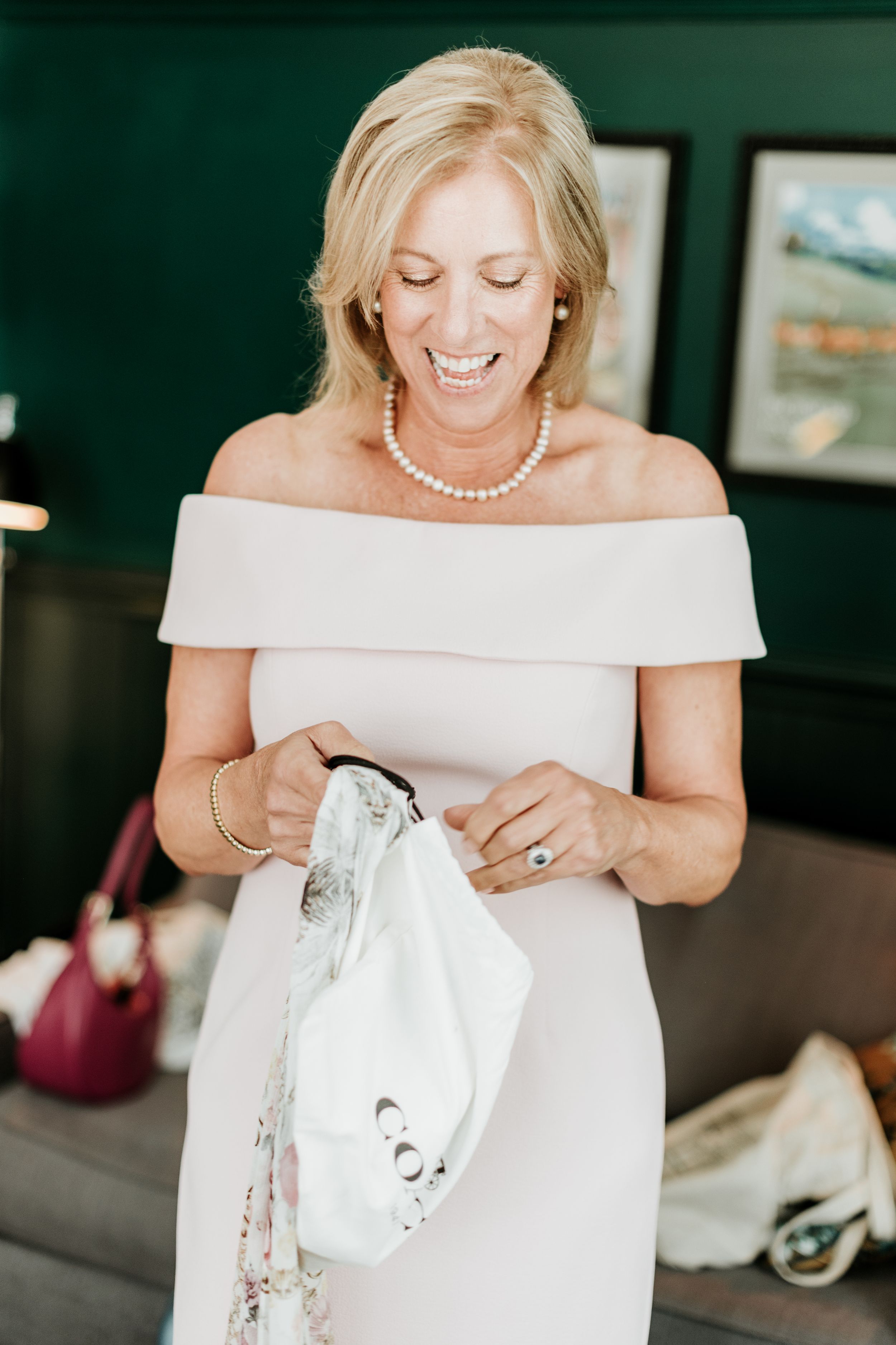 A bride in an elegant off-shoulder white dress and pearl necklace smiles while getting ready.