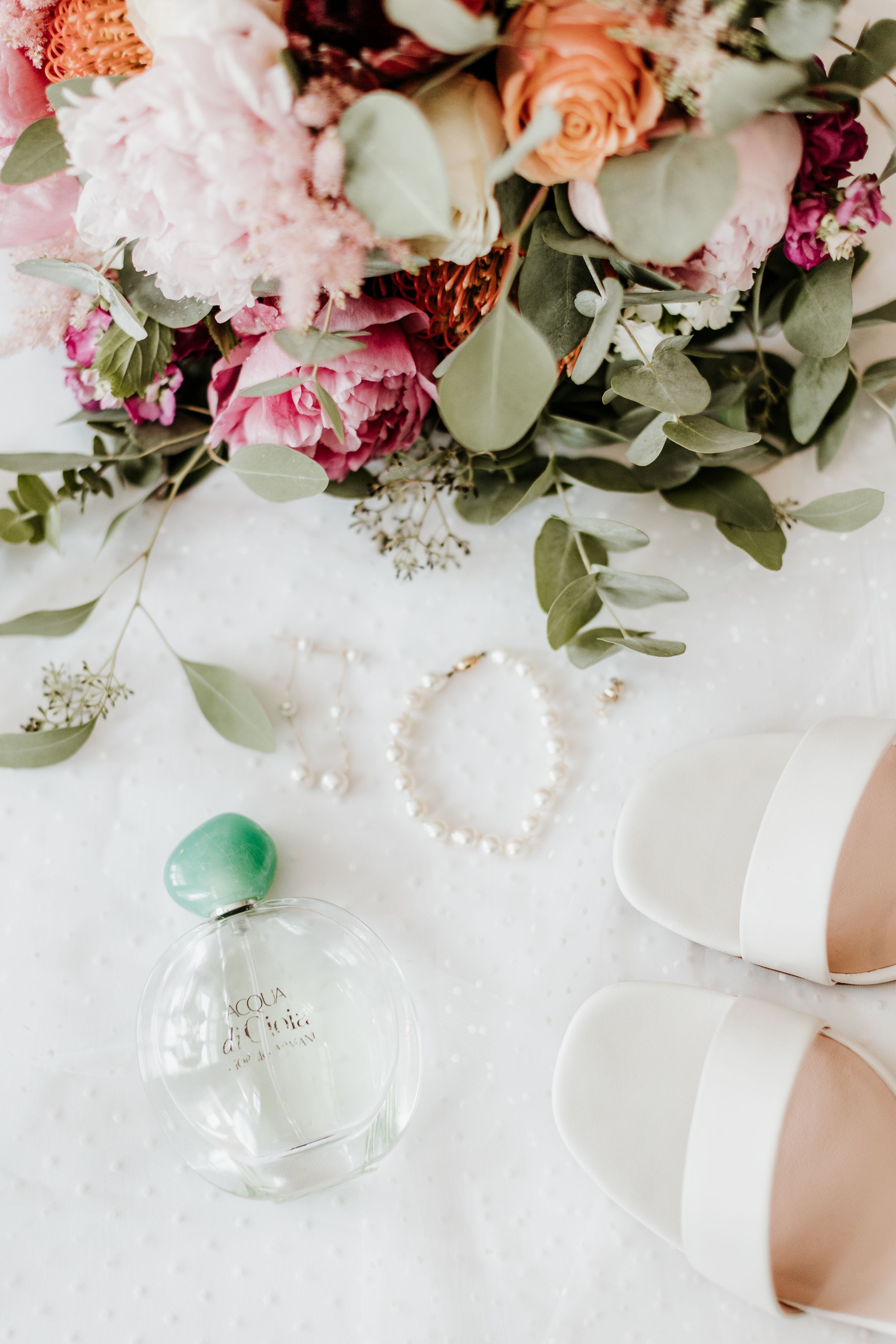 Elegant wedding details including pink and peach floral arrangements, a pearl bracelet, perfume bottle and white bridal shoes.