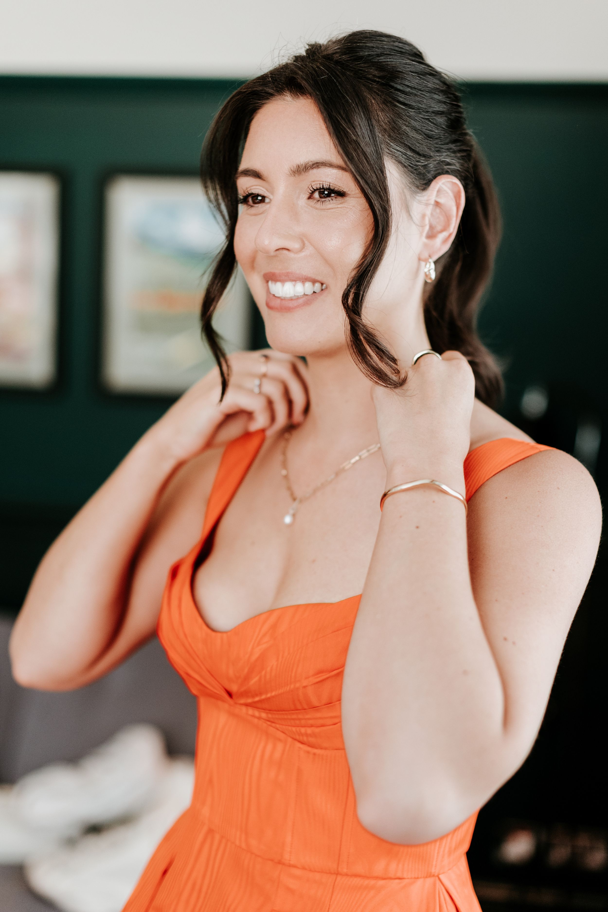 A woman in a vibrant orange dress smiles while adjusting her earring.