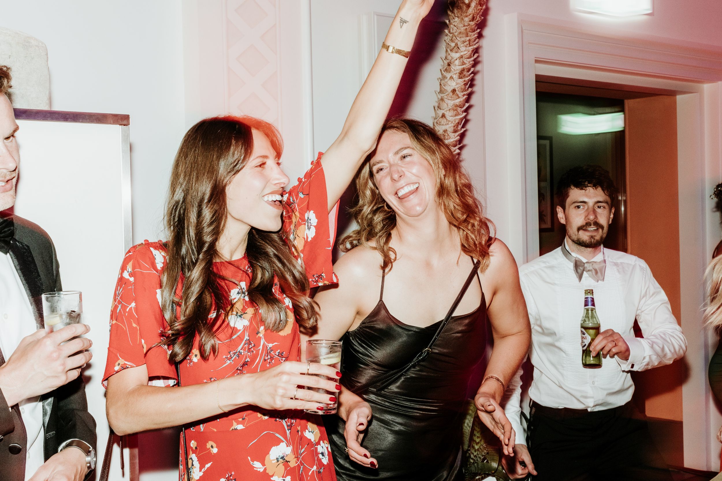 A group of friends laugh together at a party, with one wearing a red floral dress and another in a black satin slip dress.