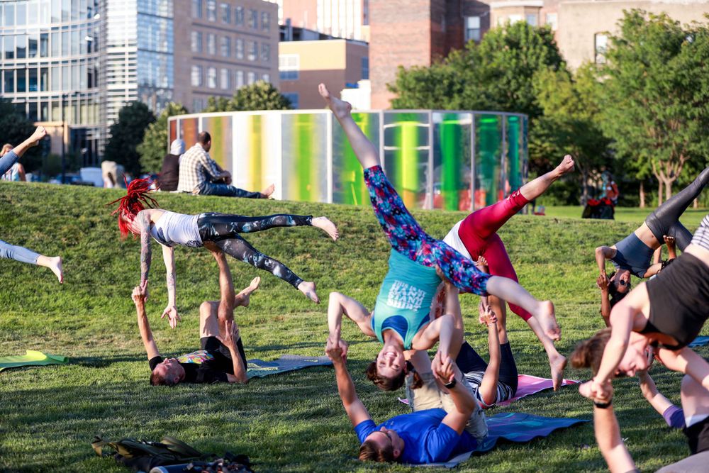 People practice acrobatic yoga poses on green grass in an urban park on a sunny day.
