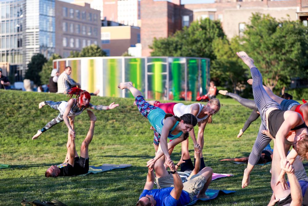 People practicing acrobatic yoga and partner balancing poses in a grassy urban park on a sunny day.