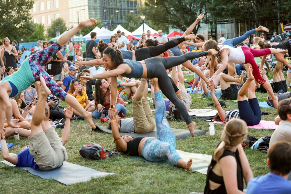 Group of people practicing partner acrobatic yoga poses on grass during an outdoor fitness event in a city park.