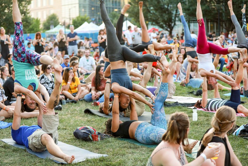 Large group practicing various acrobatic yoga poses together on grass during an outdoor wellness festival in the park.