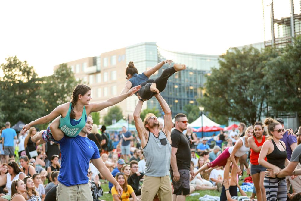 Dancers perform acrobatic lifts at an outdoor summer festival while spectators watch in the background.