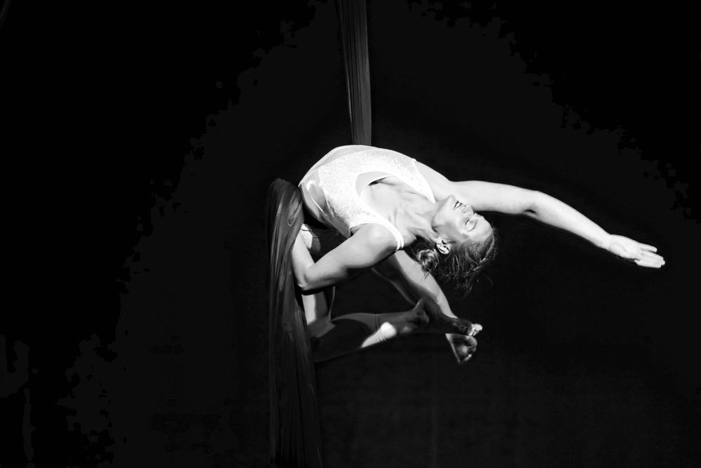 A performer in white costume executes an aerial acrobatic move on a pole against a dark background in dramatic lighting.