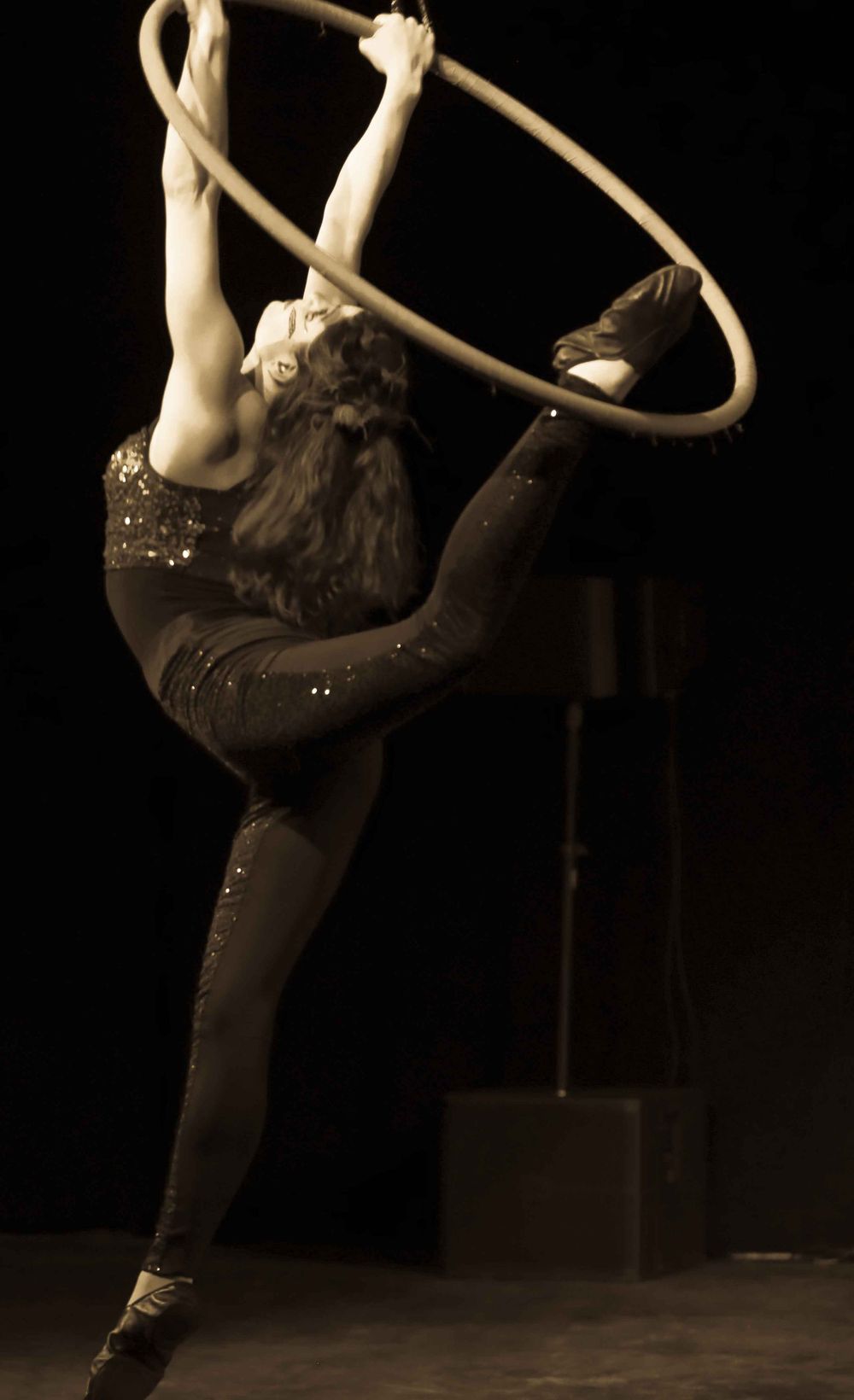 Performer in a dark sparkly costume demonstrates aerial acrobatics with a hoop on stage in sepia tone lighting.