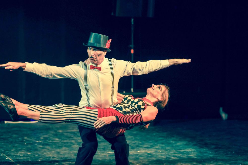 Two circus performers in vintage costumes demonstrate a balanced acrobatic pose during a stage performance.