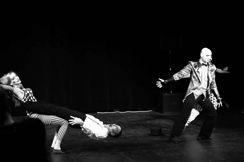 Three performers in black and white theatrical costumes create a dramatic scene on a dark stage during a performance.
