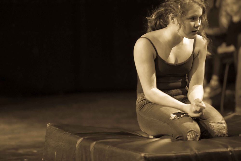 Performer in dark dress sits contemplatively on stage in sepia-toned theatrical performance.