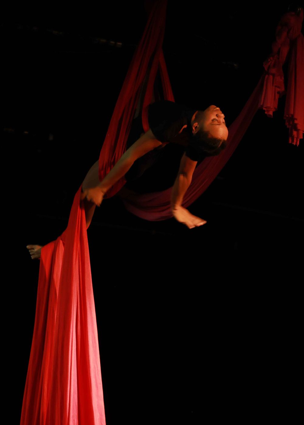 A performer in dark attire gracefully poses while suspended from red aerial silks against a black background.