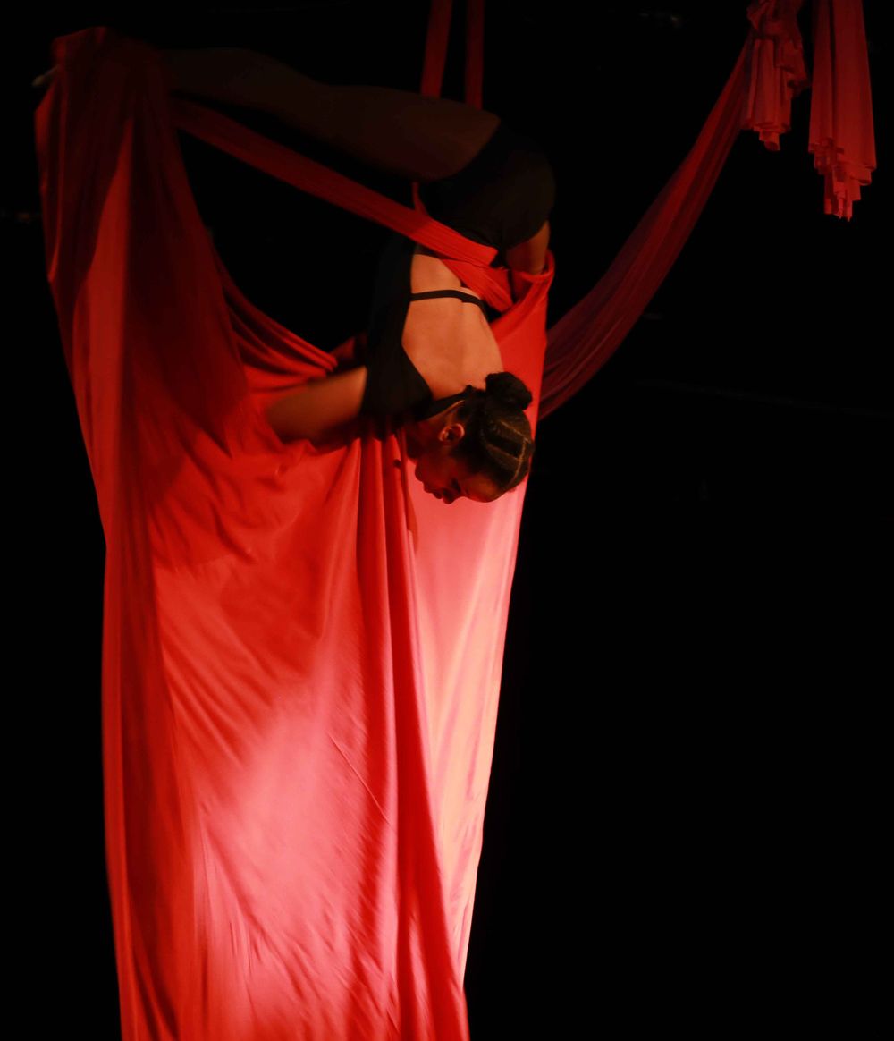 A silhouetted performer gracefully suspends in red aerial silk against a dark backdrop with dramatic lighting.