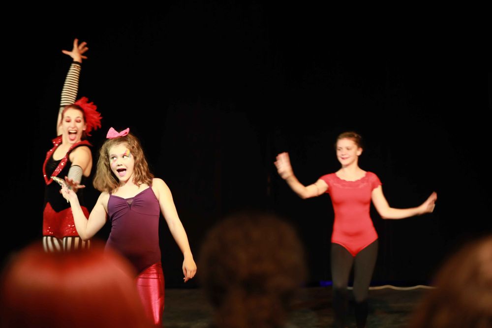 Dancers in burgundy and red leotards perform an energetic routine on a dark stage during a dance recital.
