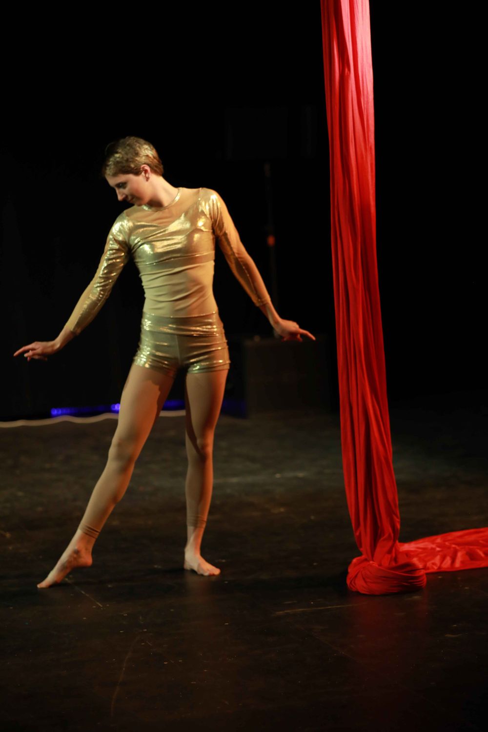 Dancer in gold metallic bodysuit performs on dark stage next to flowing red silk aerial fabric.