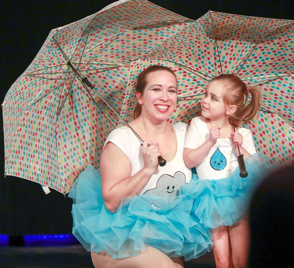 Two figures in matching blue tutus pose together beneath a colorful polka dot umbrella during a performance.