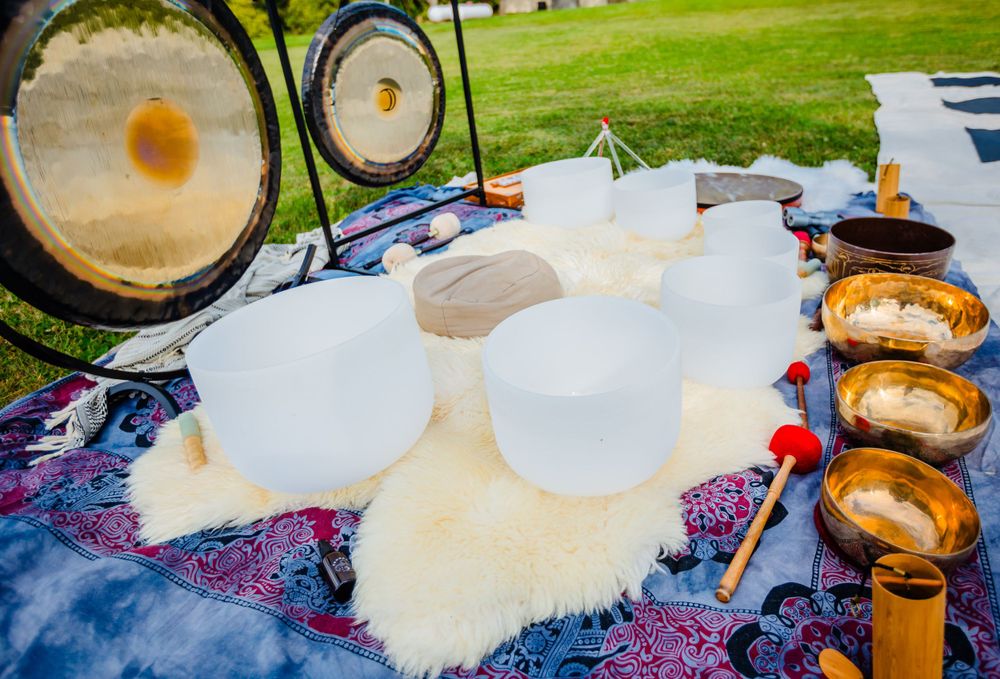 Crystal singing bowls and meditation cushions arranged on a colorful blanket with gongs in an outdoor setting.