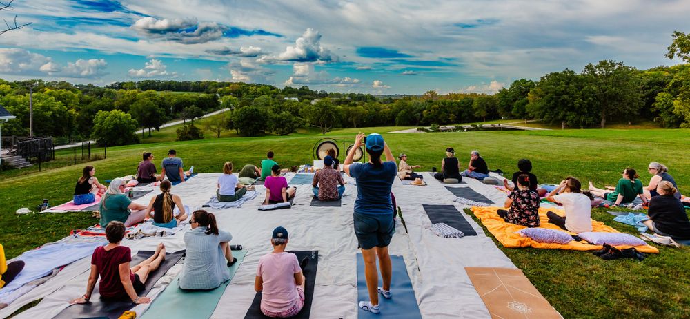 People sitting on picnic blankets enjoying an outdoor event on a grassy hillside under blue skies.