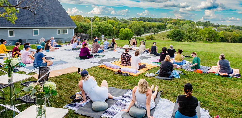 People gathered on blankets for an outdoor wellness and meditation retreat on a grassy hillside on a sunny day.