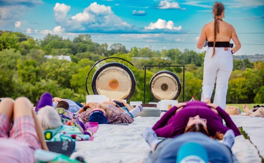 Attendees relax on blankets during an outdoor sound healing session with gongs in a scenic natural setting.