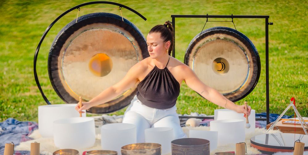 A wellness practitioner performing crystal bowl sound healing with singing bowls and gongs in a sunny outdoor setting.
