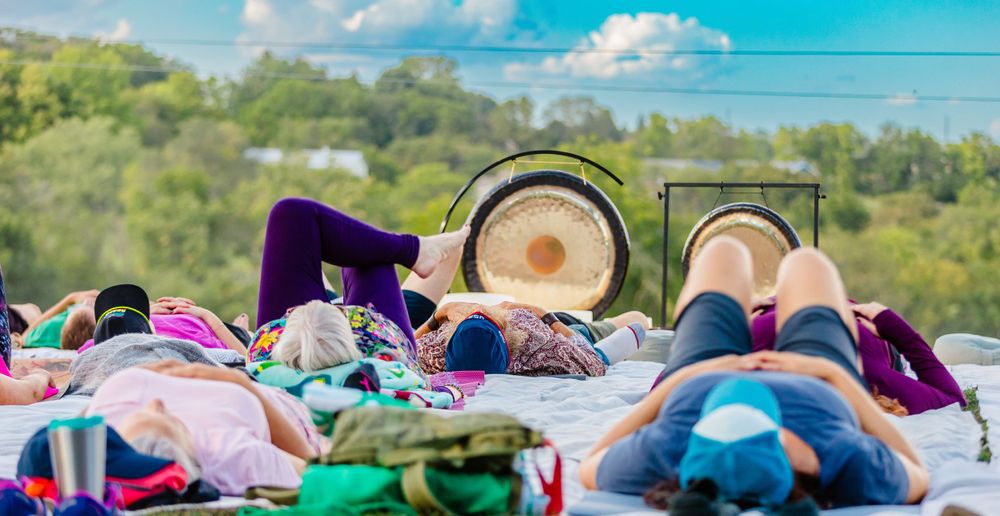 Colorful yoga mats and meditation cushions arranged outdoors with gongs in the background against a bright blue sky.