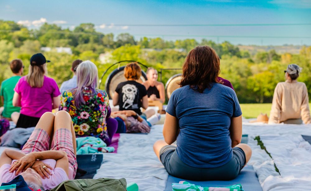 A group gathers outdoors for a meditation session on a hilltop overlooking lush greenery and blue skies.