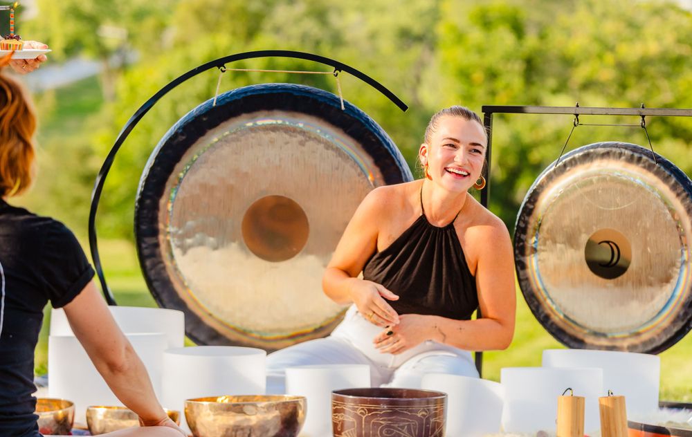 Sound healing practitioner sits by large meditation gongs during an outdoor wellness session.