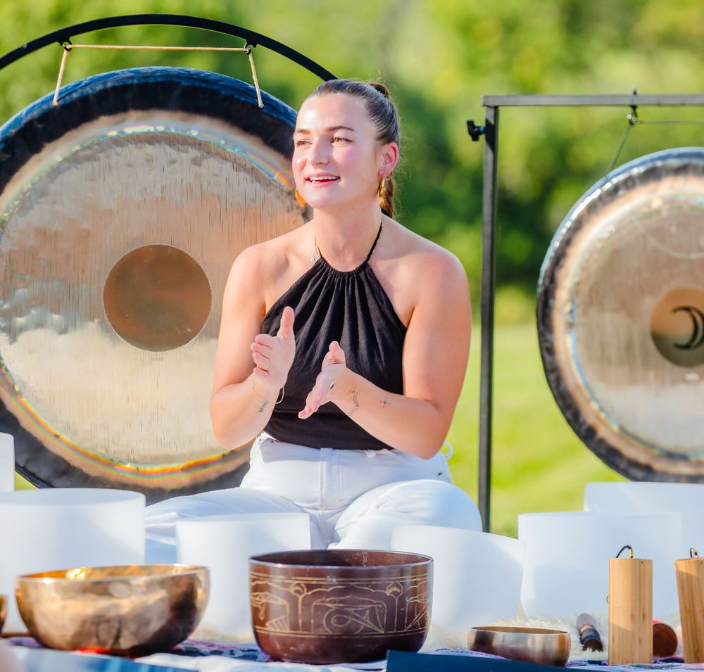 Practitioner sits cross-legged with meditation bowls and gongs during an outdoor sound healing session.