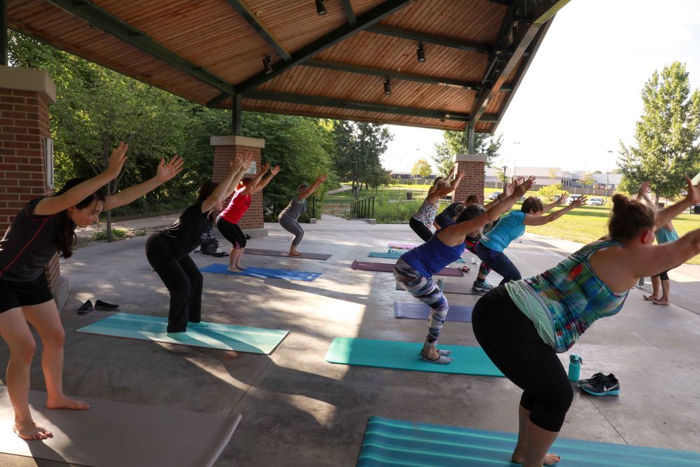A group of people participate in an outdoor fitness class doing stretching exercises under a covered pavilion.